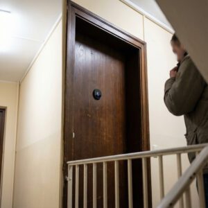 Ein verunsicherter Bewohner blickt im schmalen Flur auf eine glänzende Kameralinse in einer dunklen Holztür.